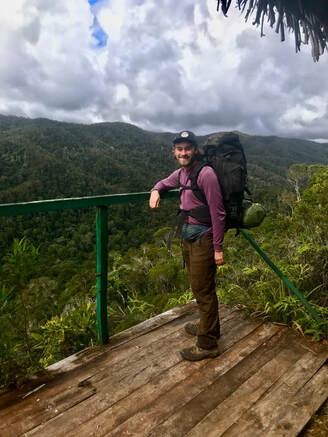 Eric stands on a platform wearing a backpack and smiling at the camera with forested hills and clouds in the background.
