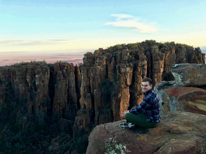 Photo of Eric Wuesthoff sitting on the edge of a cliff and smiling with rock formations in the background.