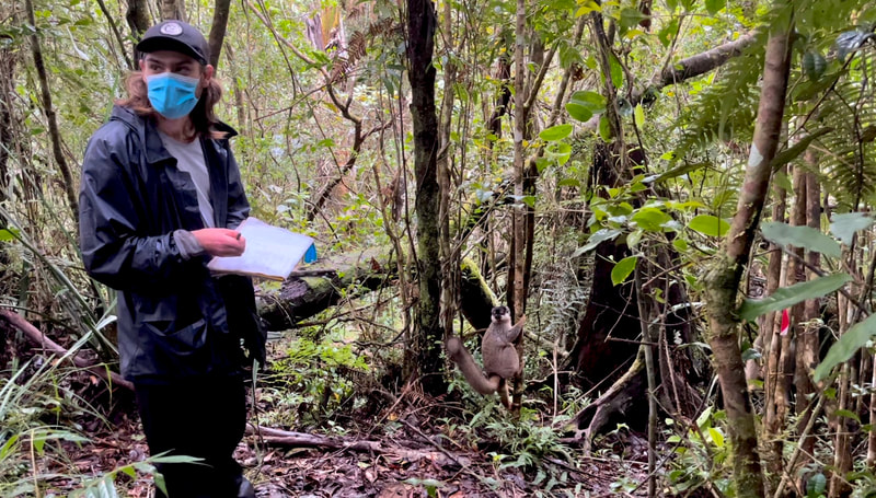 Photo of Eric Wuesthoff masked and holding a pen and notebook with trees and a lemur in the background.