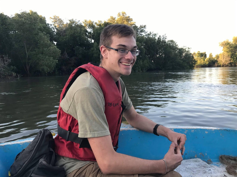 Photo of Eric Wuesthoff smiling and wearing a life jacket while sitting in a boat with a river and trees in the background.