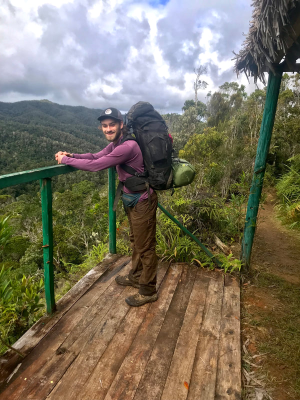 Photo of Eric Wuesthoff wearing a backpack and leaning on a railing with forested mountains in the background.