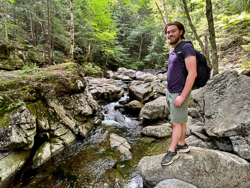 Photo of Eric Wuesthoff wearing backpack and standing on rock with stream and trees in the background.